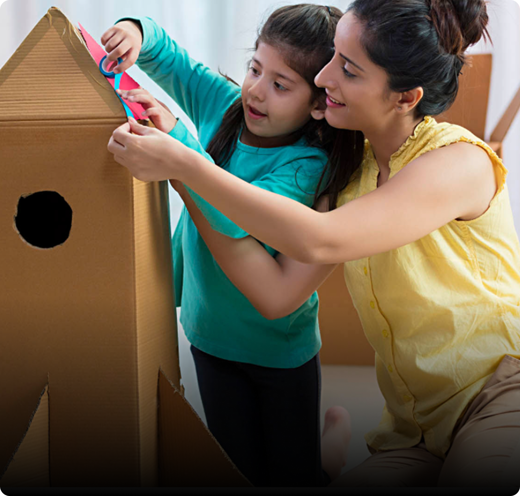 Woman and young girl decorating a cardboard playhouse together, with the girl holding pink and blue tape.